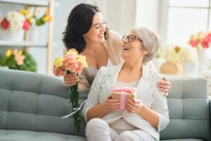 Mamma e figlia si guardano sorridenti. La mamma ha un pacco regalo in mano e la figlia ha dei fiori colorati