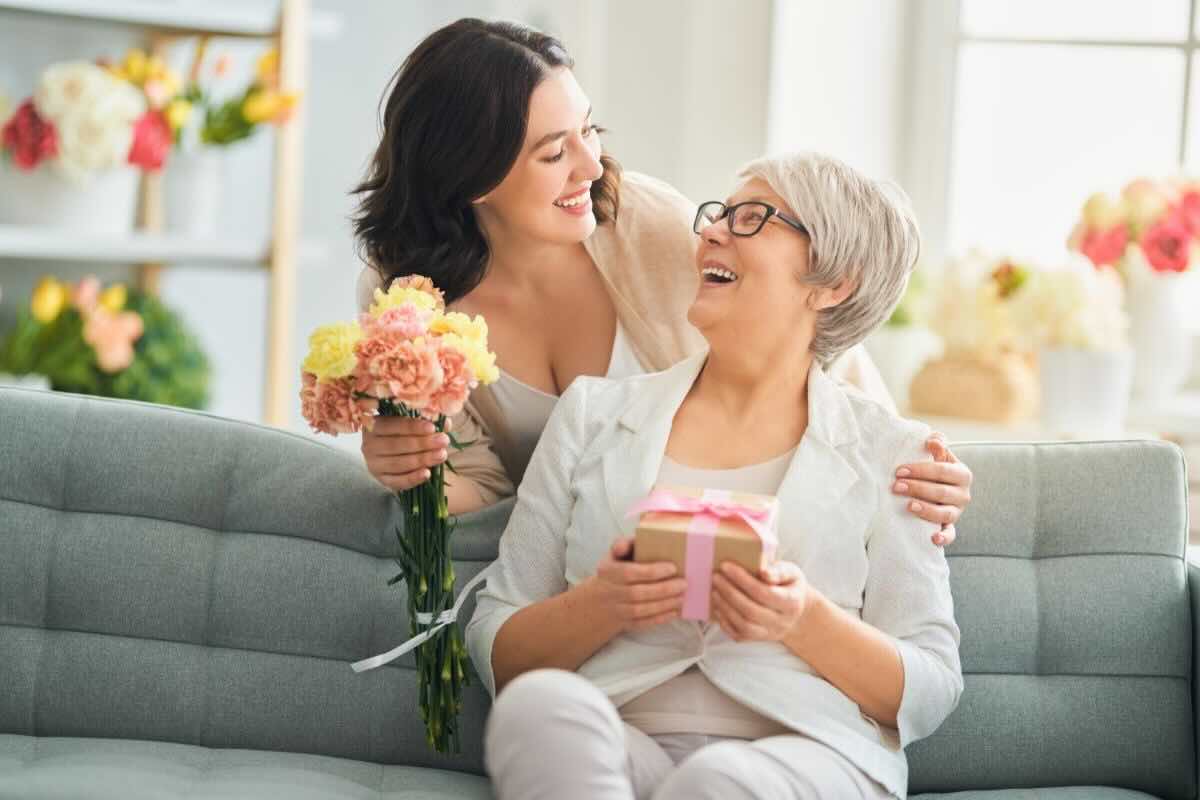 Mamma e figlia si guardano sorridenti. La mamma ha un pacco regalo in mano e la figlia ha dei fiori colorati